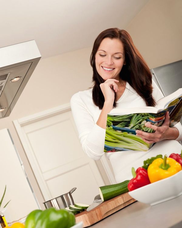 A woman in a white kitchen reads a cookbook beside a board of sweet peppers
